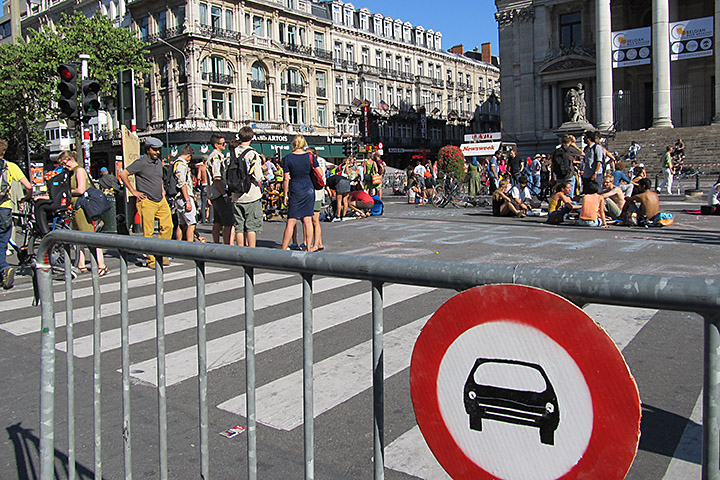 Parmaklıklı bir bariyer üzerindeki yuvarlak "Taşıt giremez" tabelası ile engellenmiş bir kent meydanında güneşli bir günde yayalar vakit geçiriyorlar.